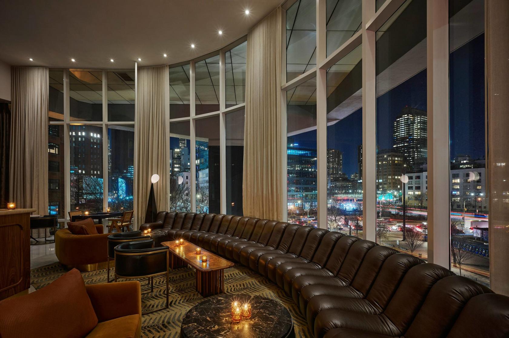 A hotel bar with floor-to-ceiling windows, city night views, curved leather seating, and candlelit tables at ModernHaus SoHo.
