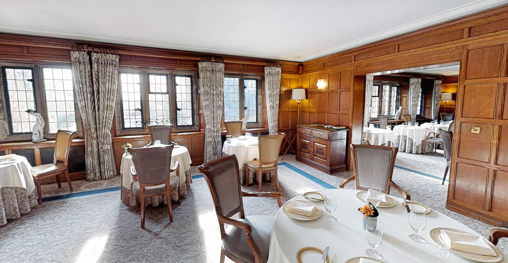 A dining room with wood-paneled walls, large windows, and round tables set with white linens at Mallory Court Country House.
