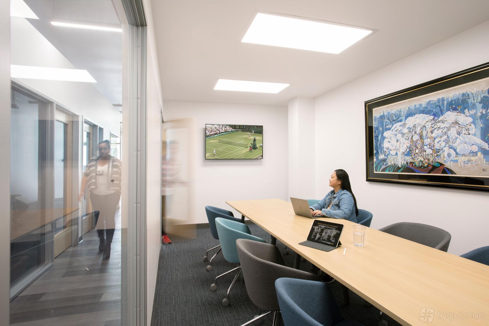 A meeting room with a long wooden table, colorful swivel chairs, wall art, and a mounted TV at BLANKSPACES Long Beach.