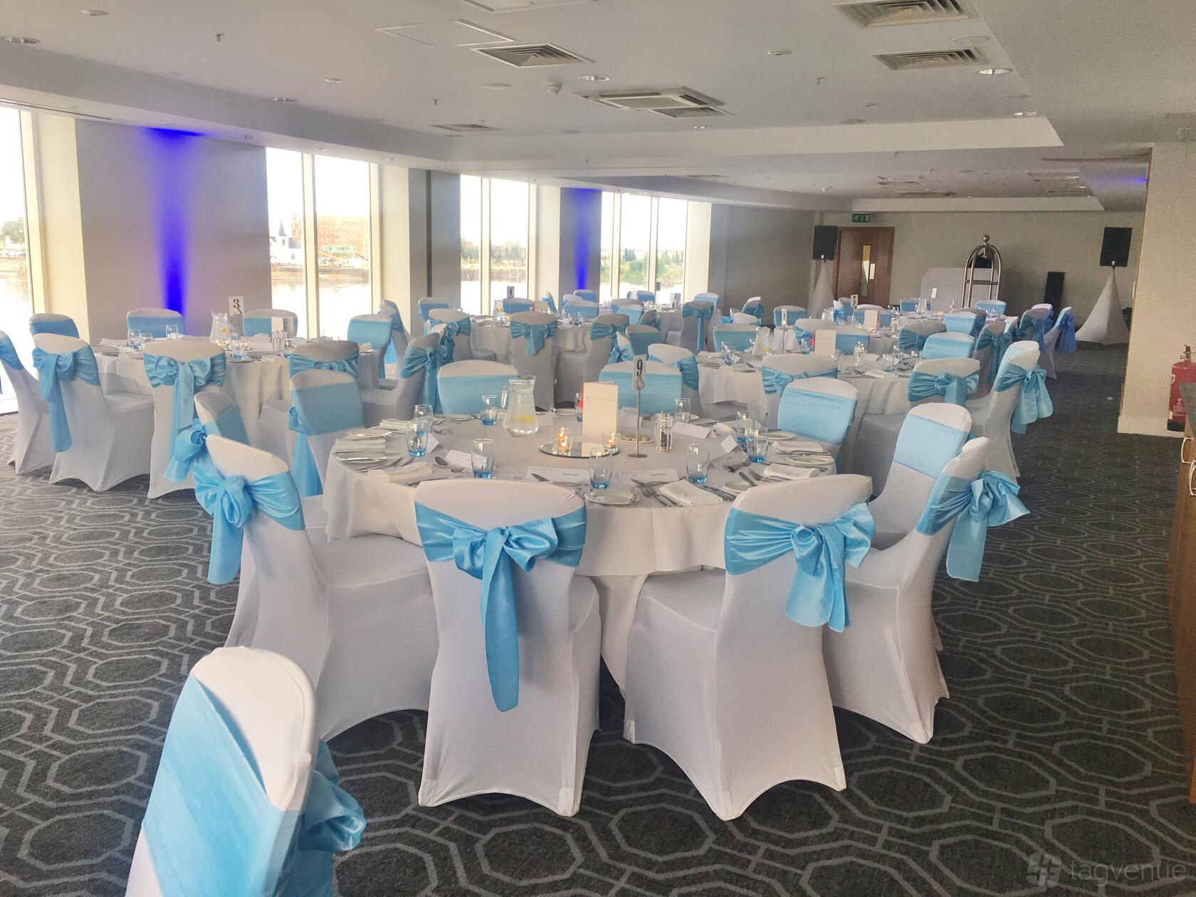 An event space with round tables draped in white linens and blue chair sashes, floor-to-ceiling windows at voco St David's Cardiff.