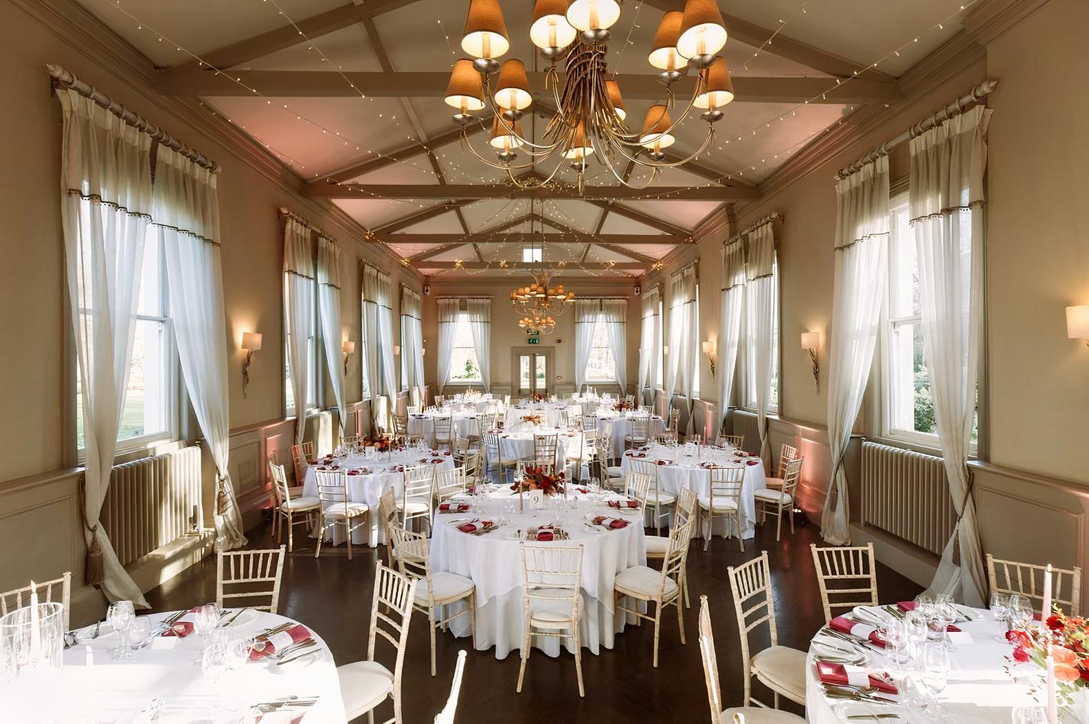 A hall with large windows, white draped curtains, chandeliers, and round tables set with white linens at Morden Hall.