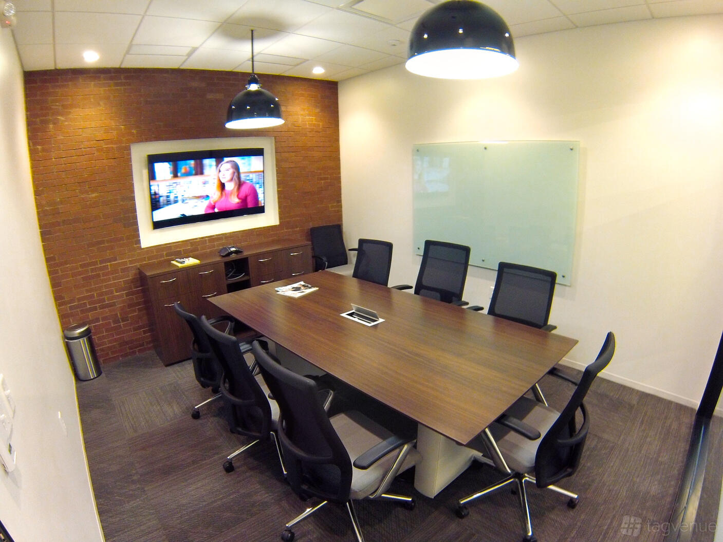 A meeting room with a rectangular wooden table, black chairs, brick accent wall, and wall-mounted TV at VenturePoint Dominion.