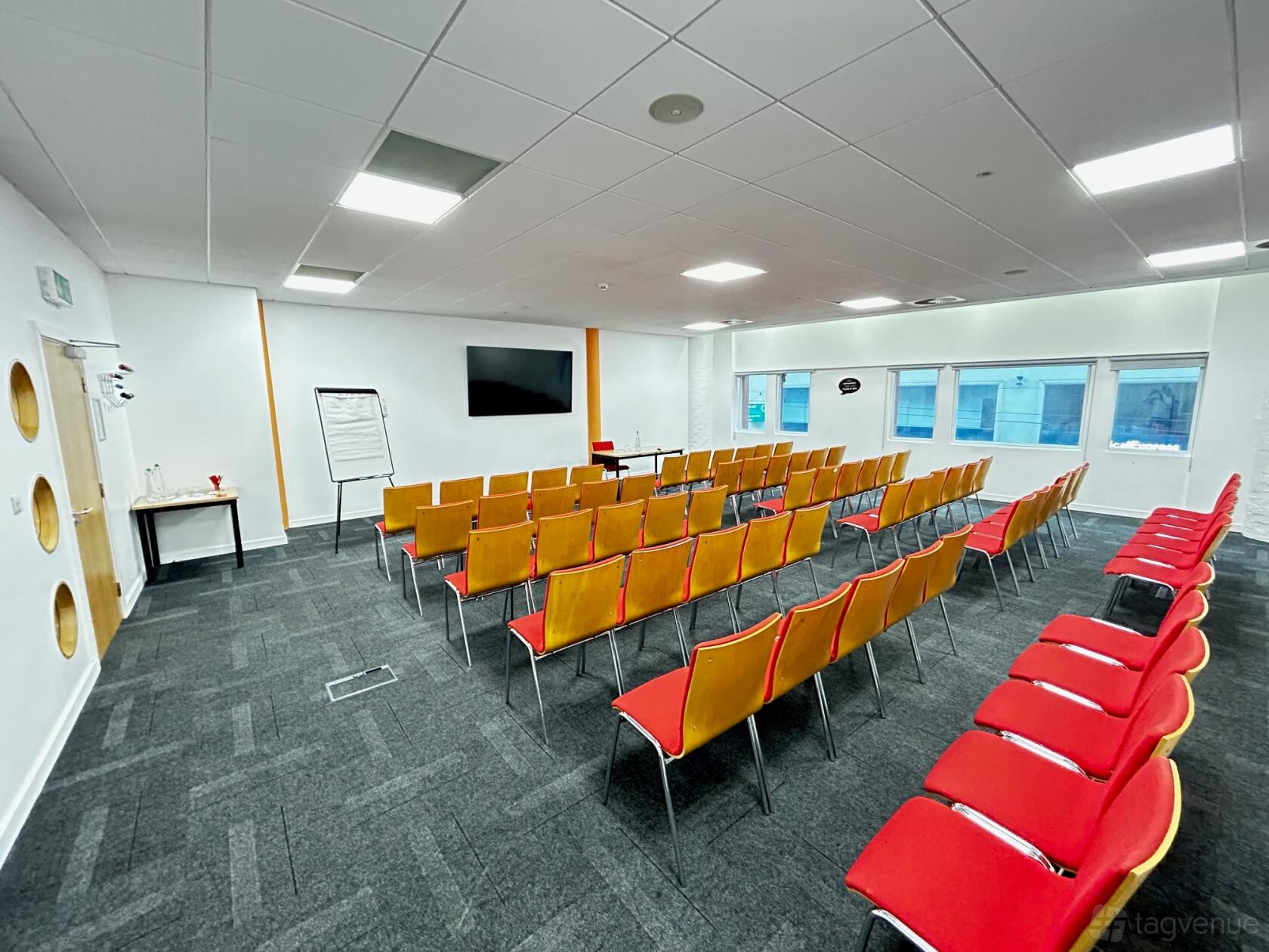A meeting centre with rows of red chairs, a wall-mounted screen, large windows, and a flipchart at The Studio Birmingham.
