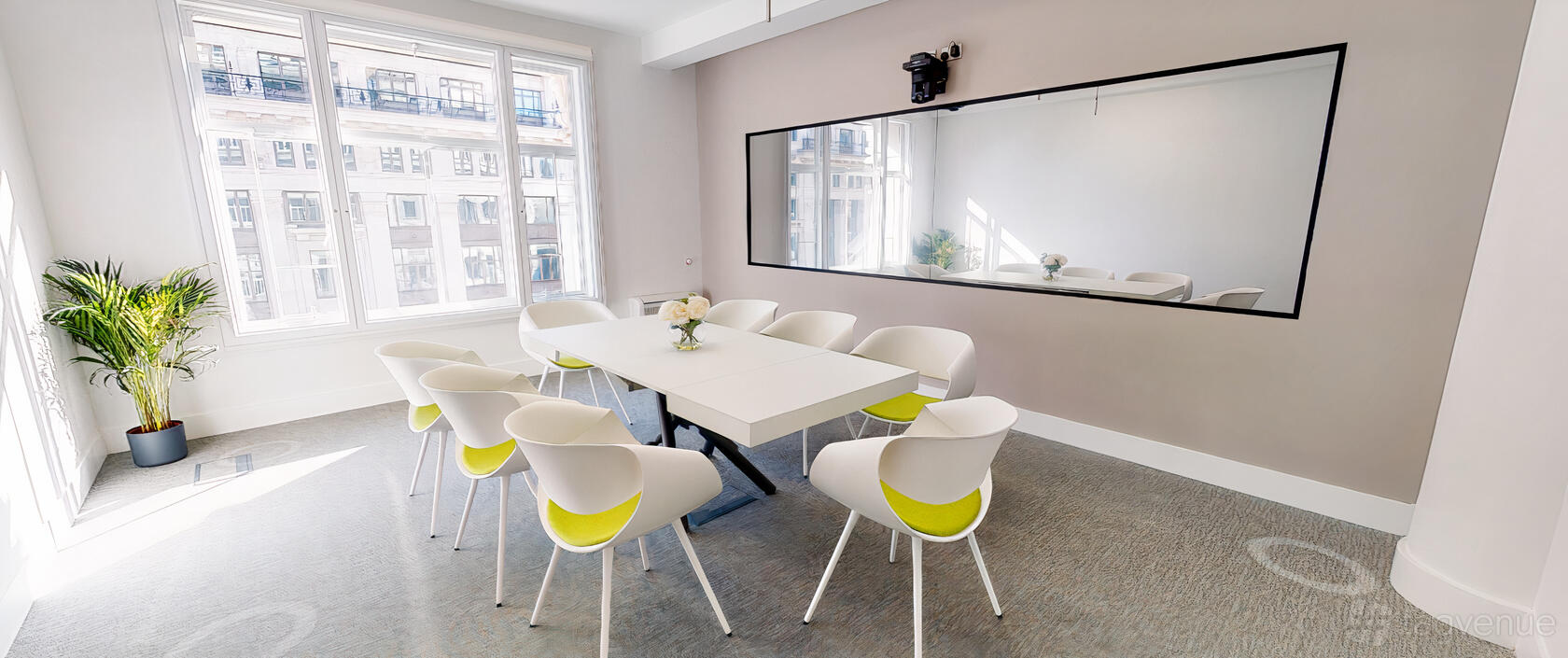 A meeting room with a long white table, white chairs with green cushions, large window, and wall mirror at SAGO Europe.
