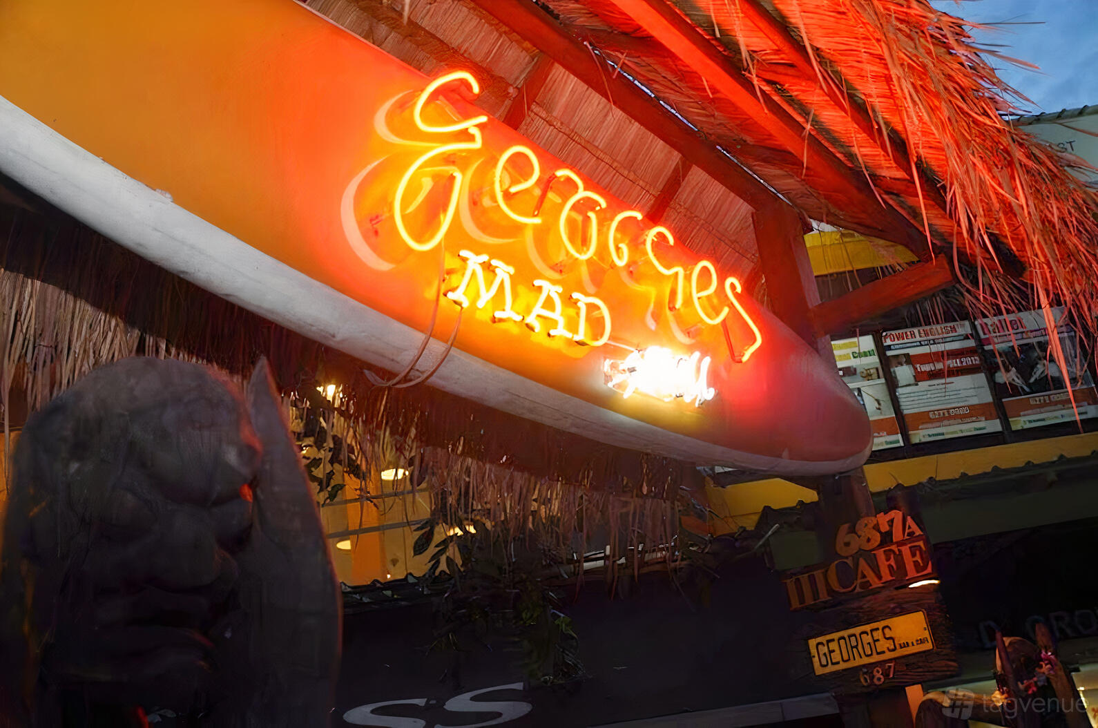 A restaurant entrance with a glowing neon sign on a surfboard and thatched roof at Georges Madbar & Grill (Siglap).