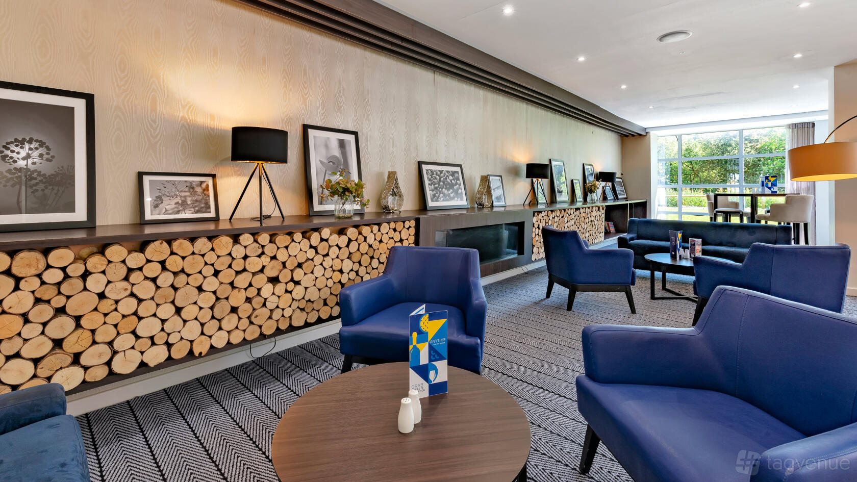 A hotel lounge with a built-in fireplace, stacks of firewood, and blue armchairs at Holiday Inn Express Leigh Sports Village.