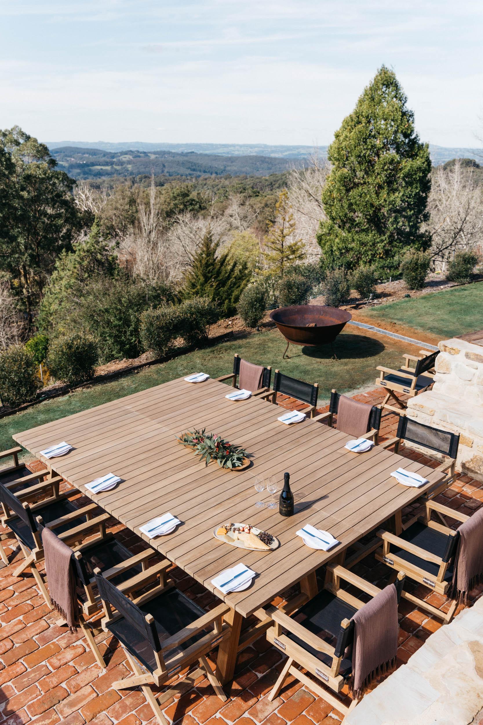 An outdoor hotel terrace with a large wooden dining table, director chairs, and a fire pit at Mount Lofty House and Estate.