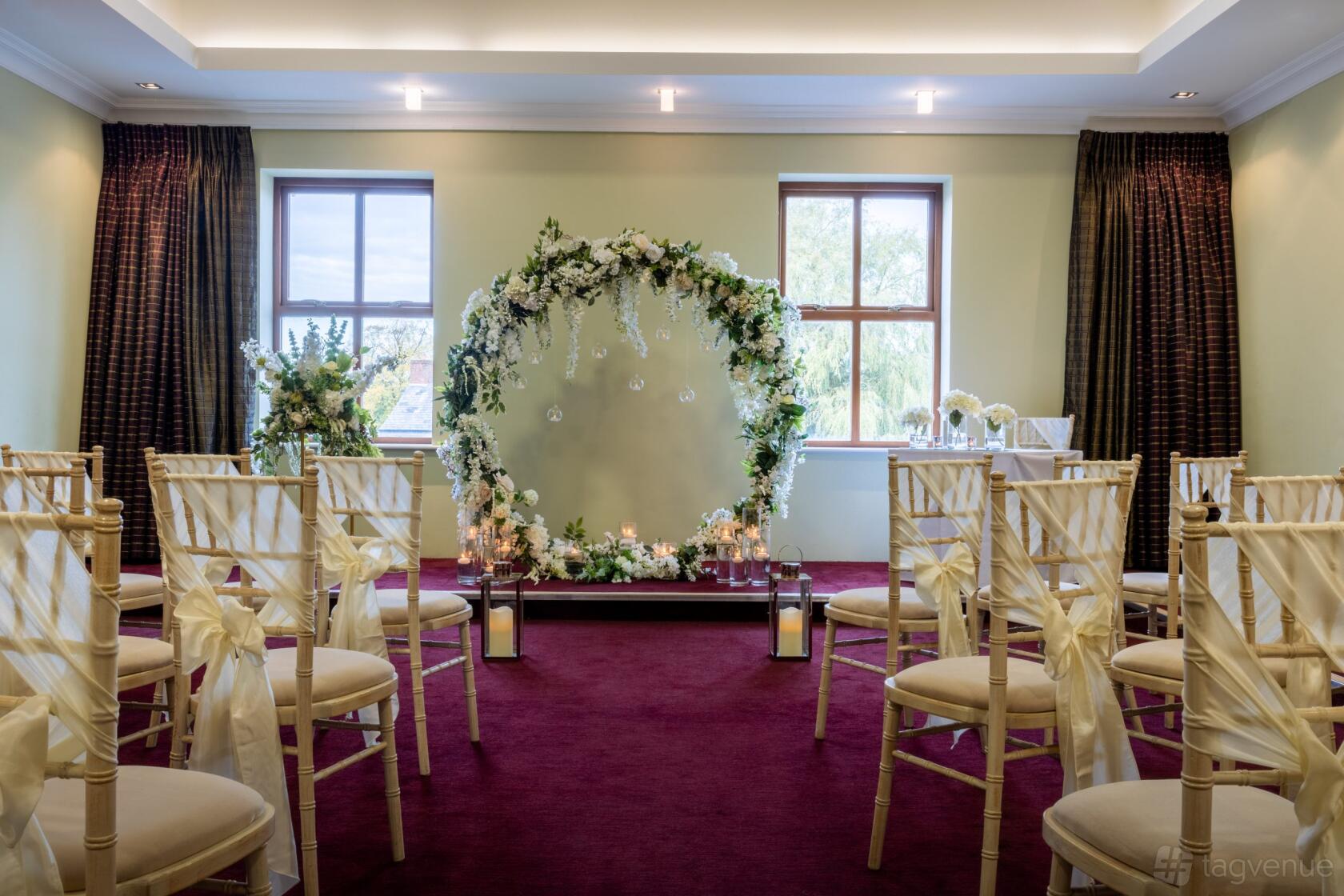 An event venue with a burgundy carpet, cream chairs with sashes, floral arch, and candle decorations at Wyrebank.