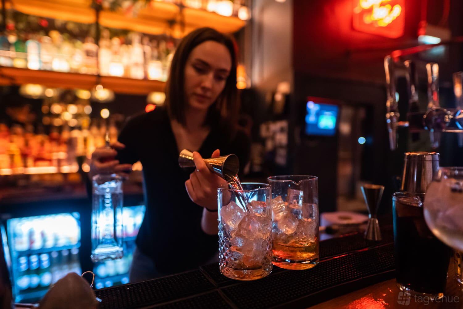 A lounge in a pub with a bartender pouring drinks over ice at a backlit bar counter at The Railway.