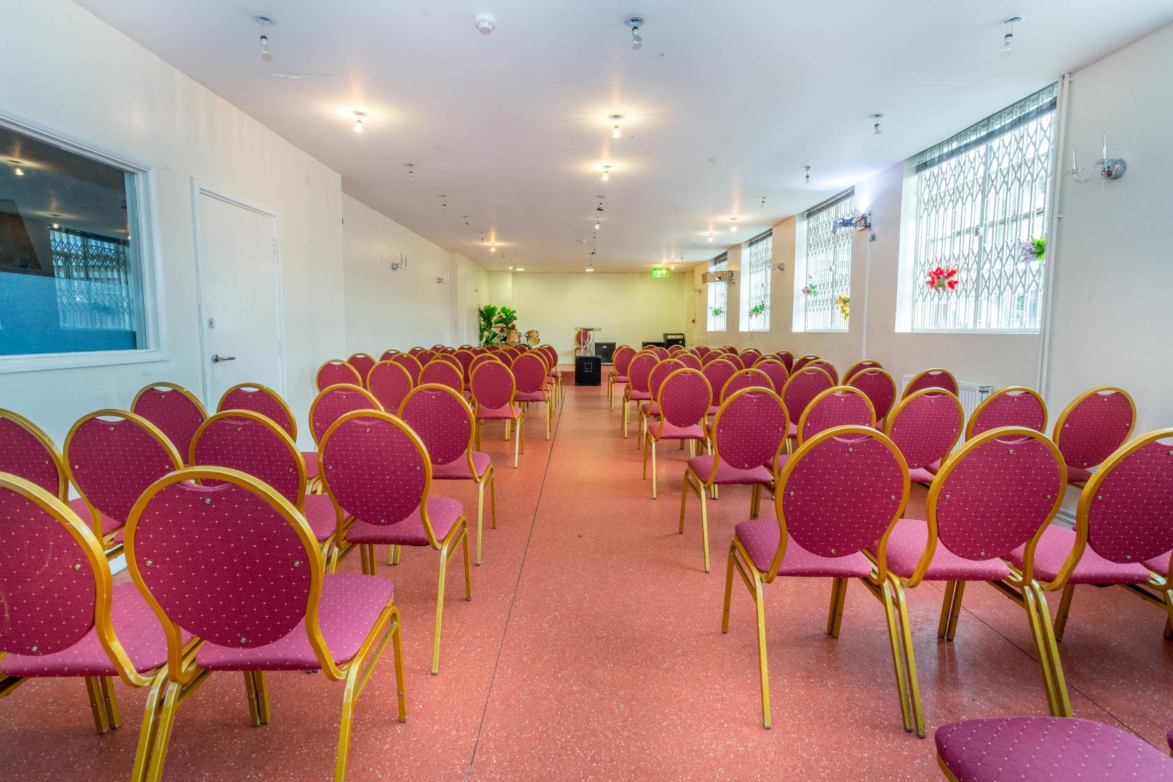 A hall with rows of red cushioned chairs, large windows with bars, and bright lighting at Excel House Halls.