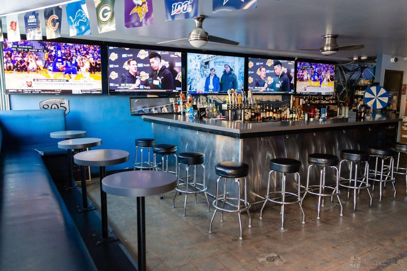 A bar with multiple flat-screen TVs, a central bar counter with barstools, and beer taps at The Blue Moon Lounge.