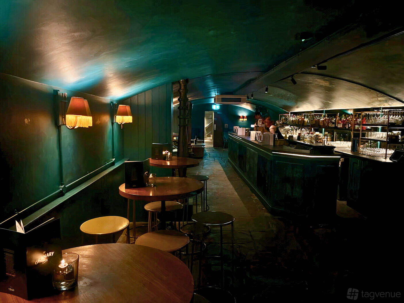 An event space in a restaurant with round wooden tables, dim lighting, and a long backlit bar at The Granary.