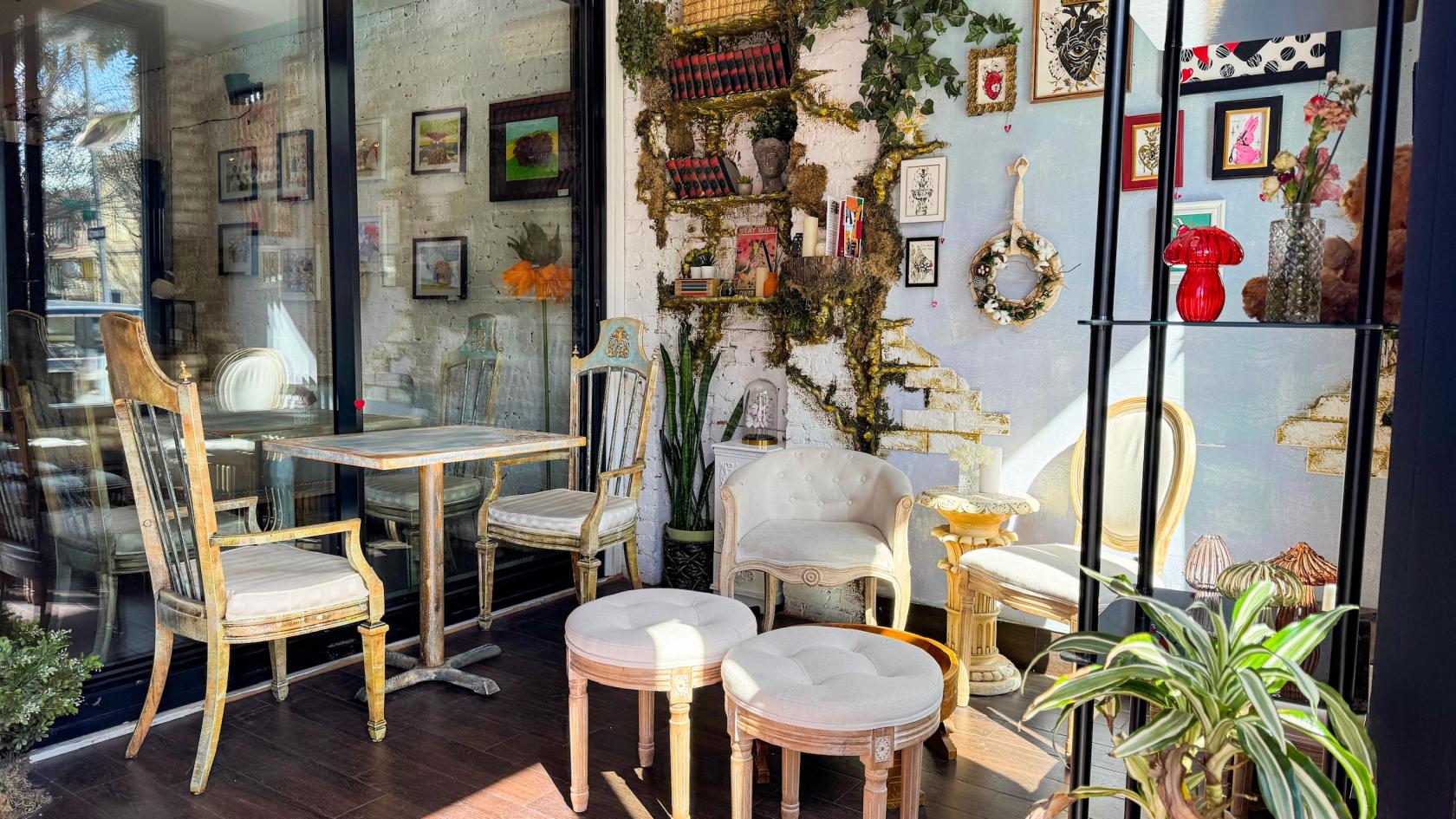 A cafe with vintage chairs, framed artwork, potted plants, and books on a brick accent wall at Whimsical, Magical Coffee & Flower Shop.