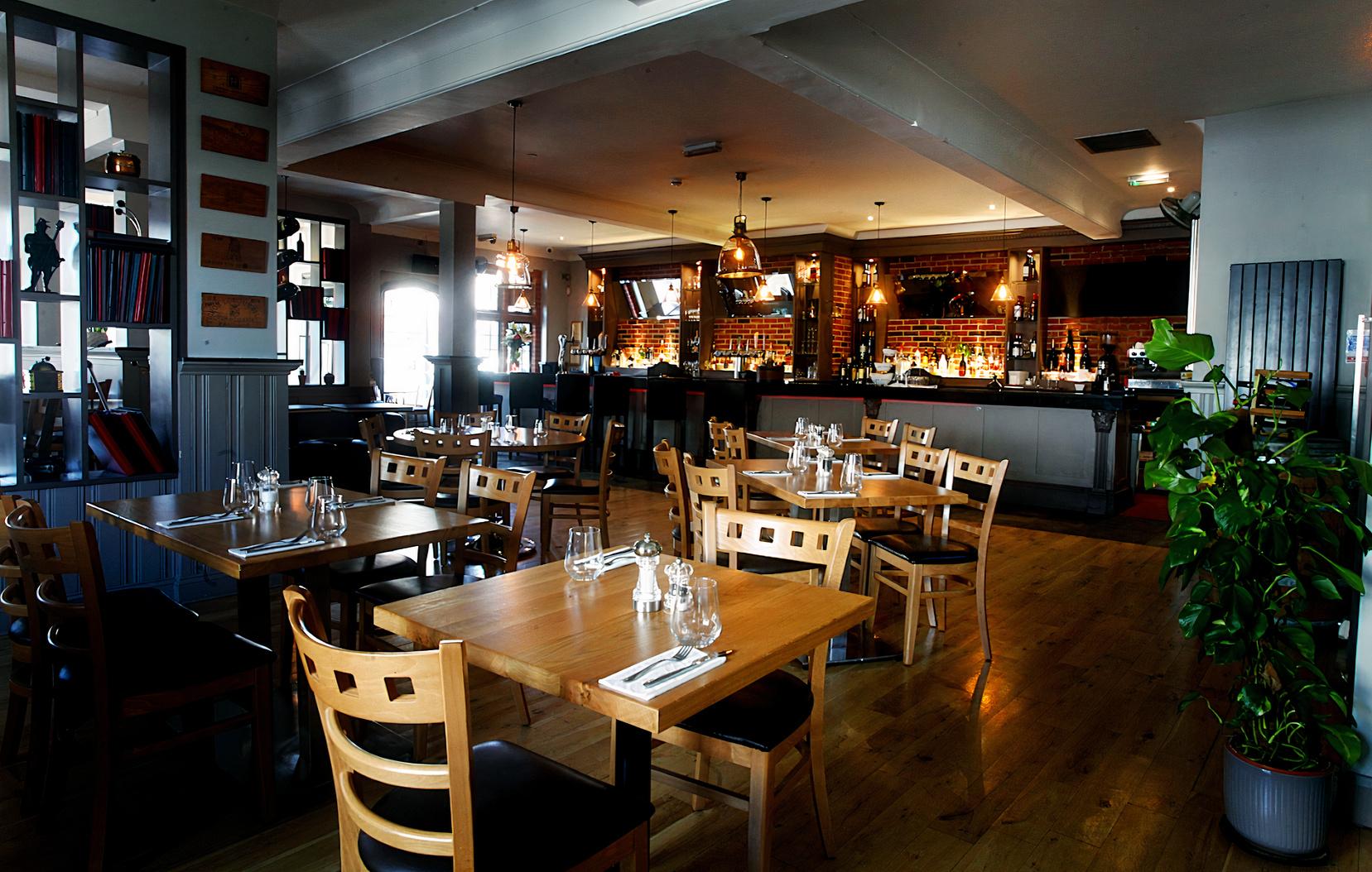 A restaurant with wooden tables, matching chairs, pendant lighting, and a brick-accented bar at Hadley House Restaurant & Bar.