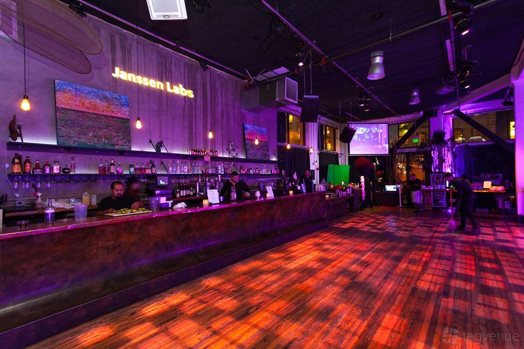 A gallery space with a long bar, wood flooring, uplighting, and abstract art on the walls at 111 Minna Gallery.