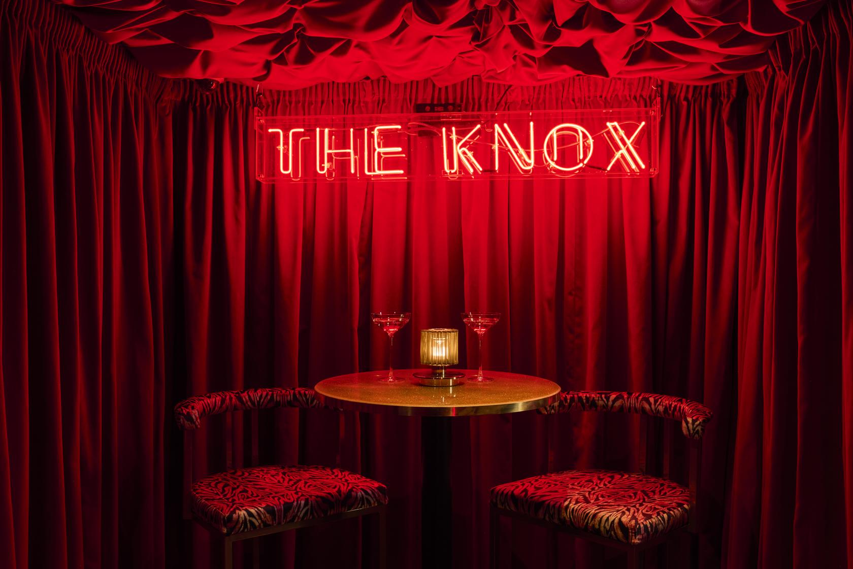 A cocktail bar lounge with a gold table, patterned stools, candle, and neon sign against red drapes at The Knox.