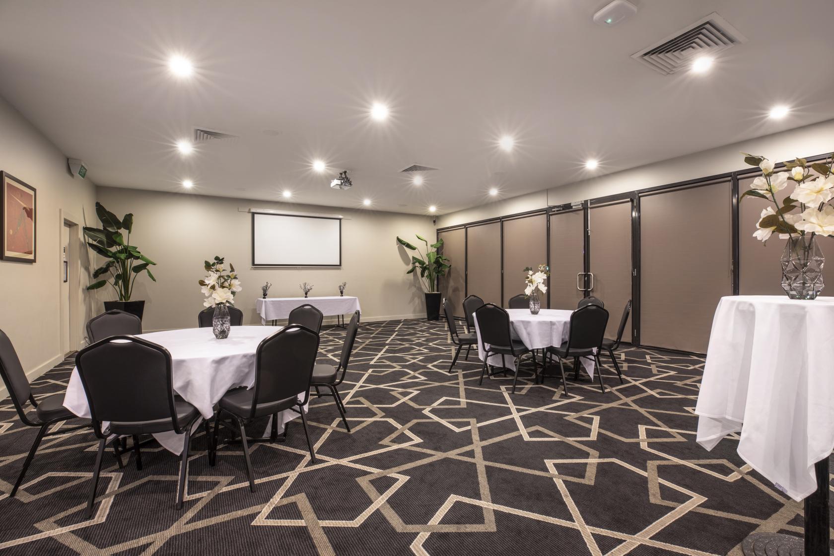 A function room in a pub with round tables draped in white linens, black chairs, and patterned carpet at Cramers Hotel.