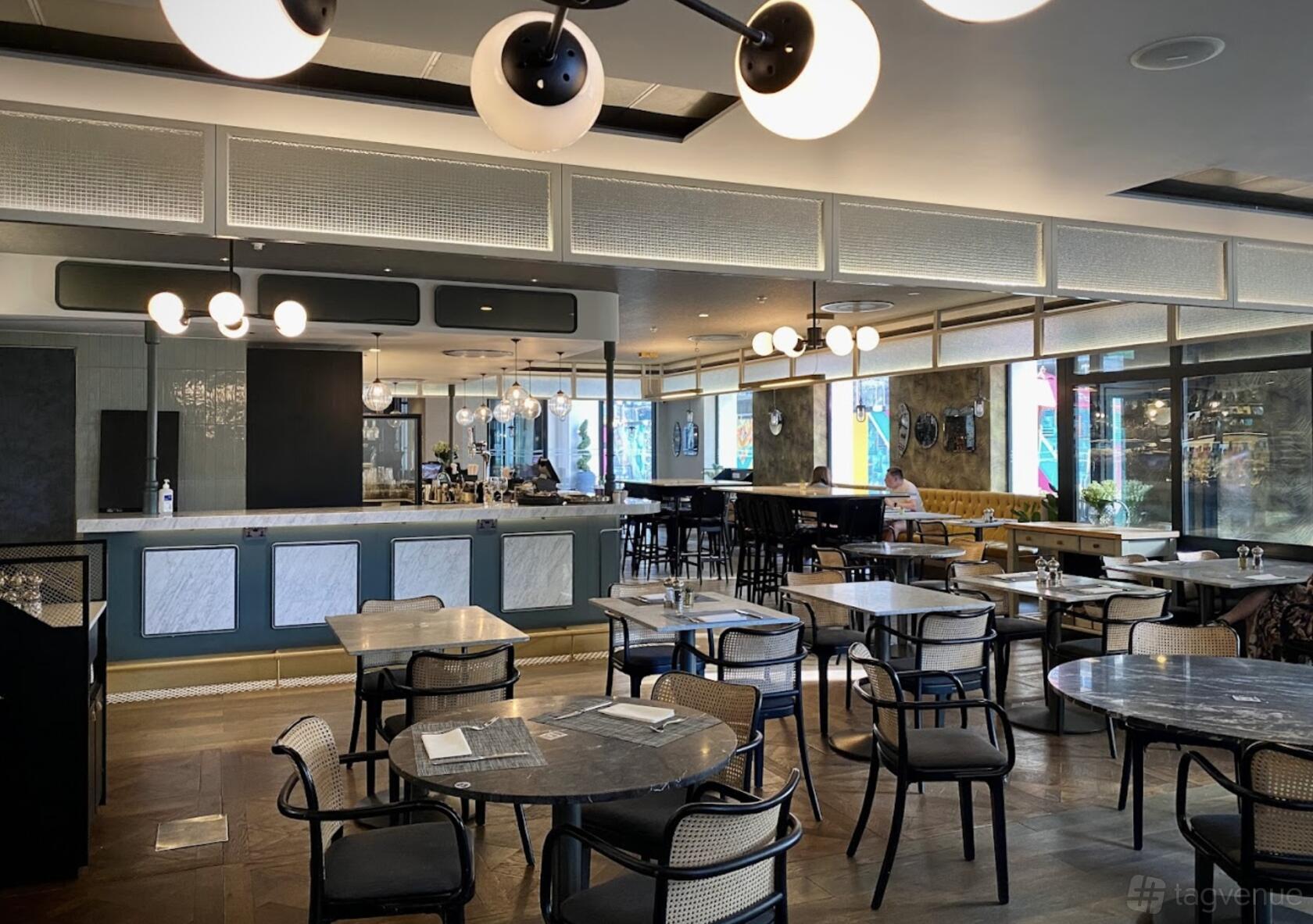 A restaurant with marble tables, wooden chairs, globe pendant lights, and large windows at Clayton Hotel City of London.