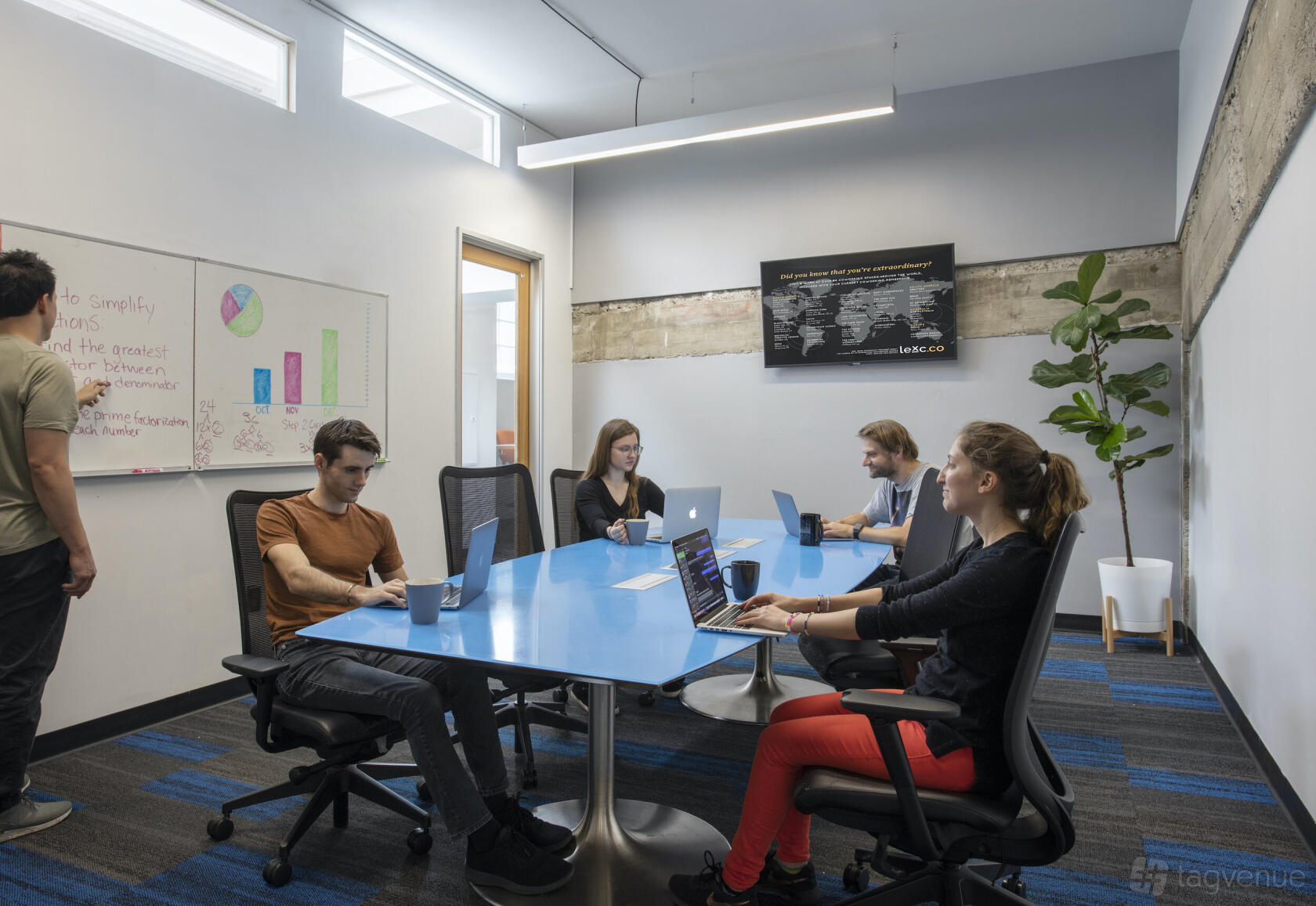 A meeting room with a blue conference table, rolling chairs, whiteboard with charts, and potted plant at BLANKSPACES Culver City.