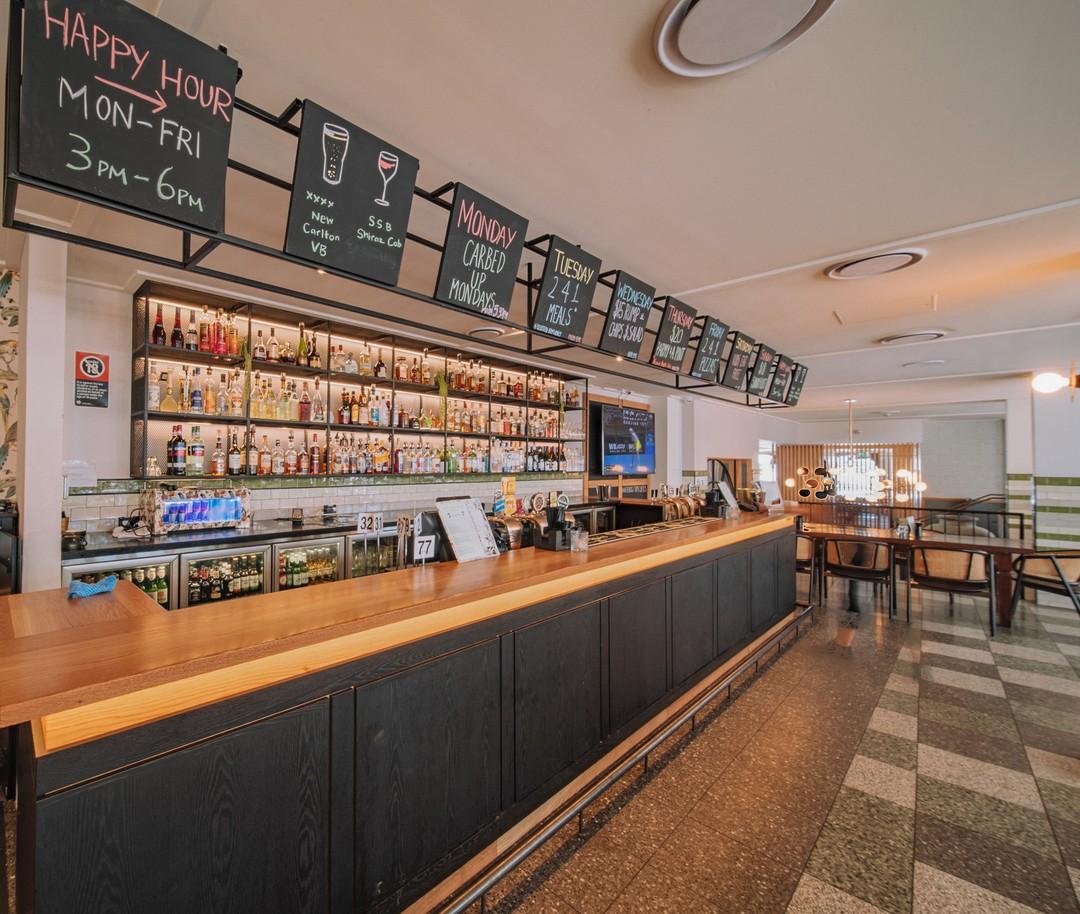 A restaurant with a long wooden bar, overhead menu boards, and a wall of shelves with bottles at Southern Cross Hotel.