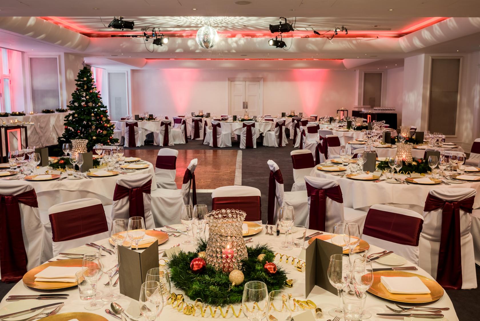 An event space with round banquet tables, burgundy chair sashes, festive centerpieces, and a decorated Christmas tree at Andaz London Hotel.