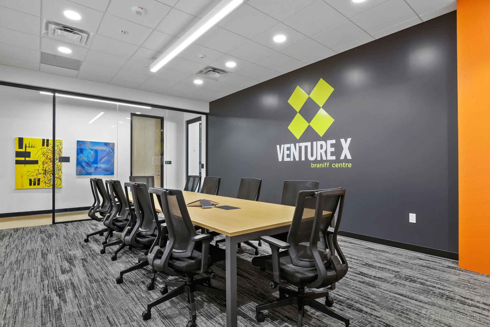 A meeting room with a long conference table, black mesh chairs, and accent wall branding at Venture X Dallas – Braniff Centre.