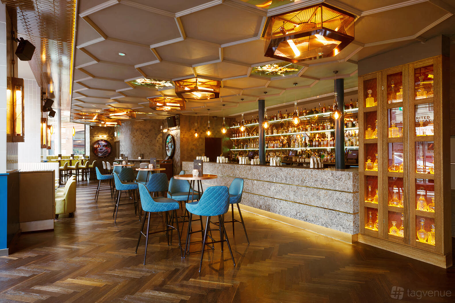 An event space in a cocktail bar with a marble bar counter, teal cushioned chairs, and geometric ceiling lights at Dirty Martini Manchester.