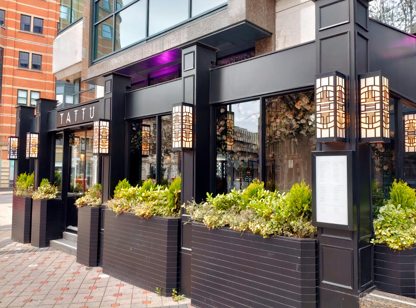 A restaurant exterior with large windows, geometric lantern lights, and flower-filled planters at TATTU Leeds.