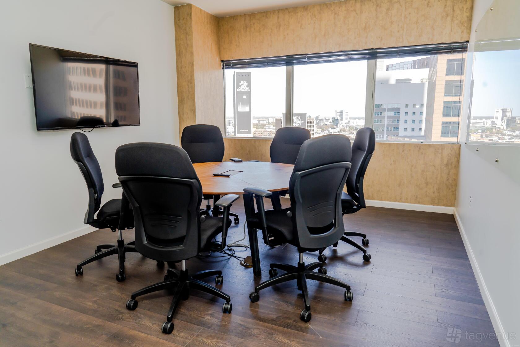 A meeting room with a round table, six black office chairs, wall-mounted screen, and city views at Connected Coworking.