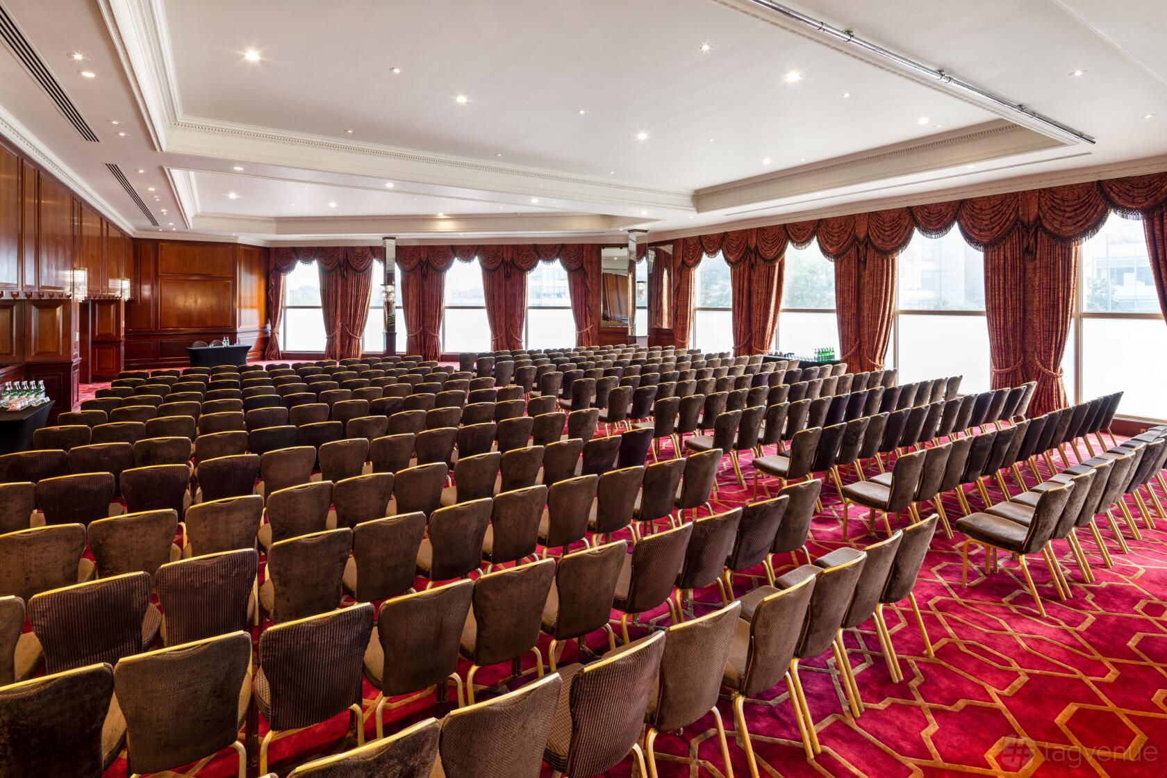 A meeting room with rows of cushioned chairs, patterned red carpet, wood panel walls, and large windows at Radisson Blu Hotel, London Heathrow.