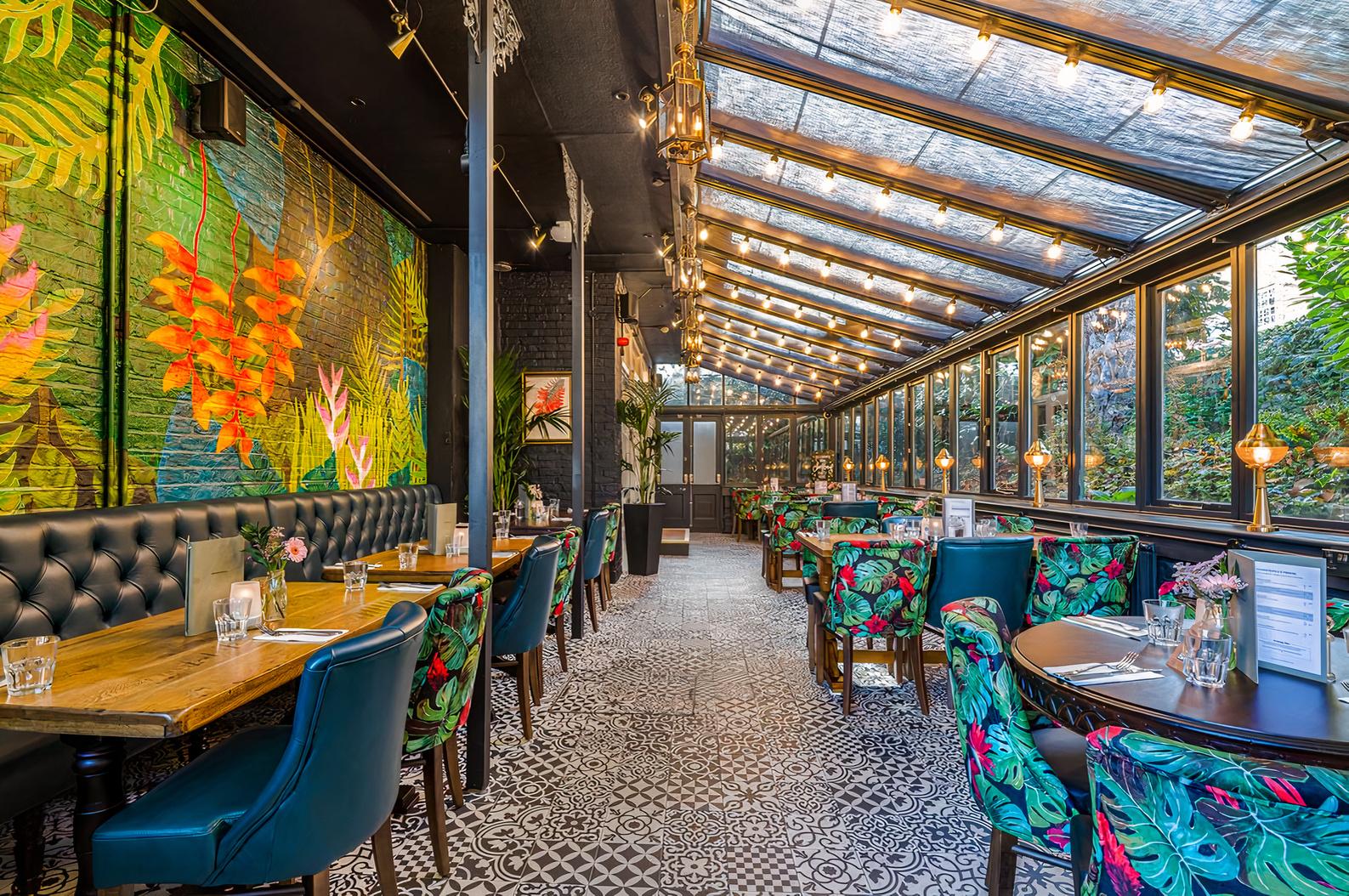 A conservatory pub space with glass roof, patterned tile floor, and botanical wall art at The Alwyne Castle.