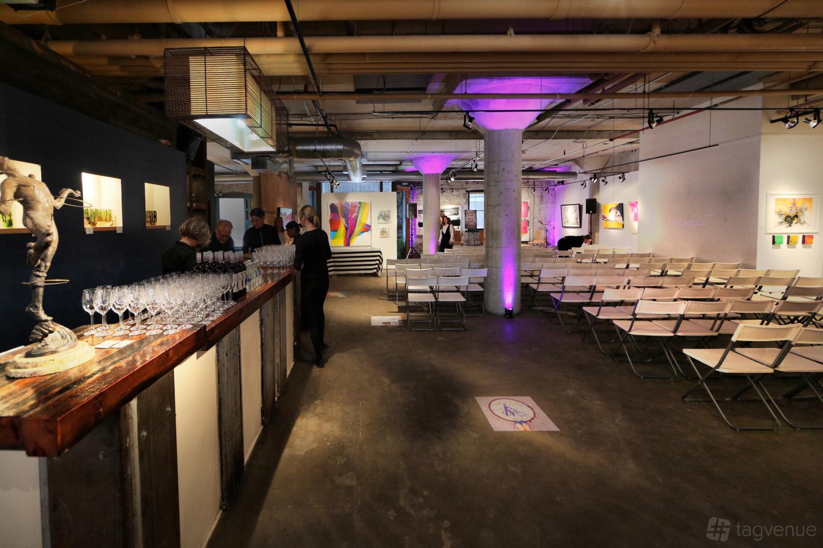 A gallery with exposed ceilings, rows of folding chairs, and modern artwork on display at Arta Gallery.