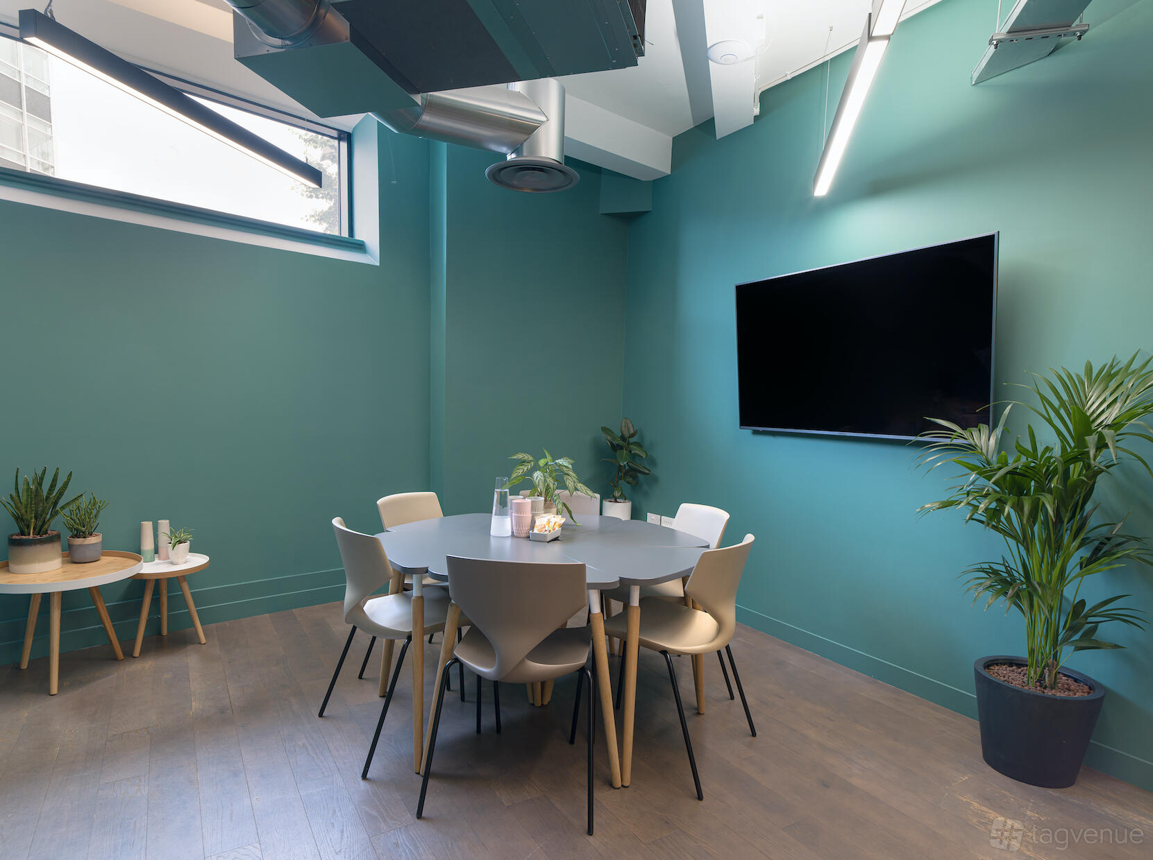 A coworking space with a round meeting table, white chairs, large wall-mounted screen, plants, and teal walls at Uncommon Borough.
