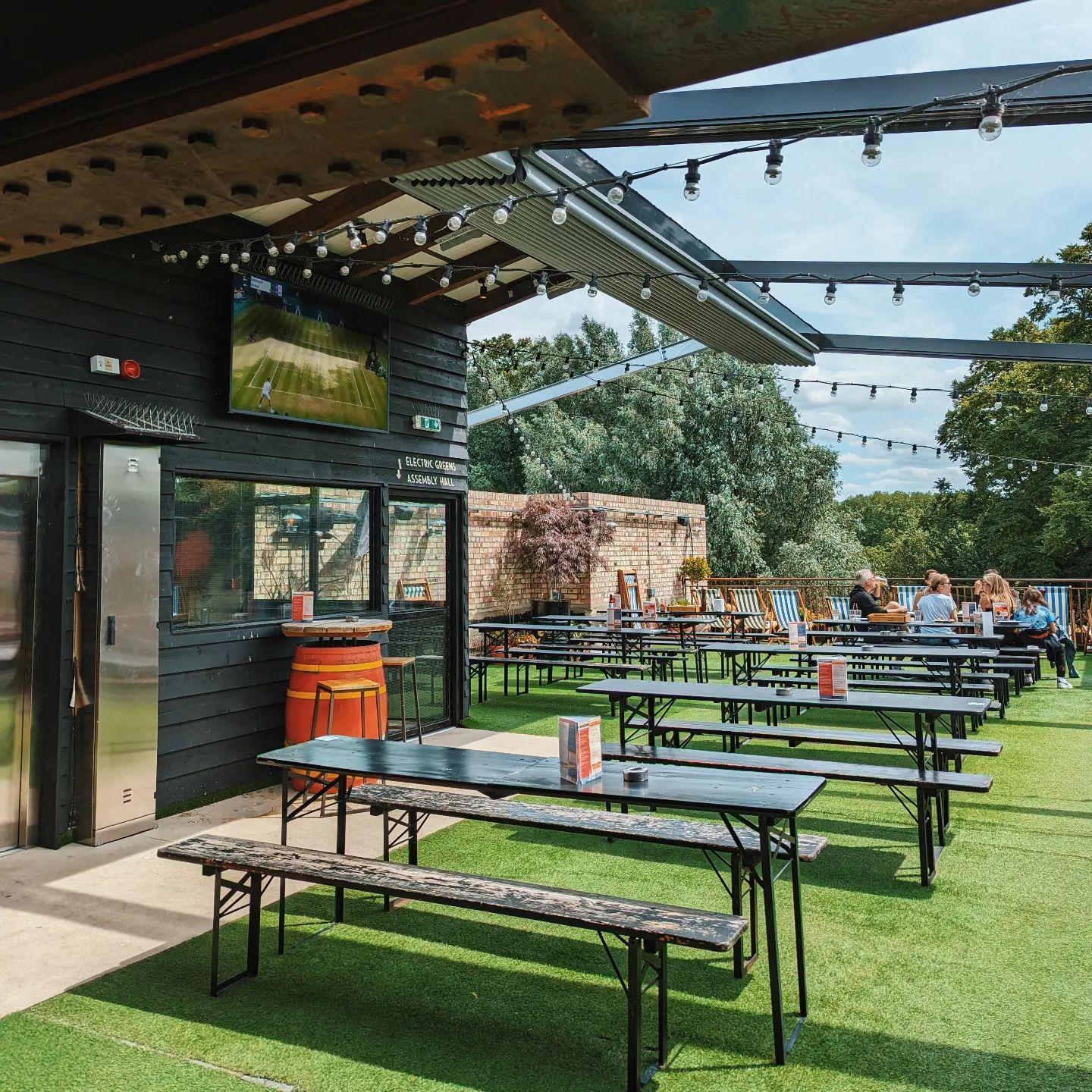 An outdoor pub area with picnic tables, artificial grass, string lights, and a TV screen at The Tivoli.