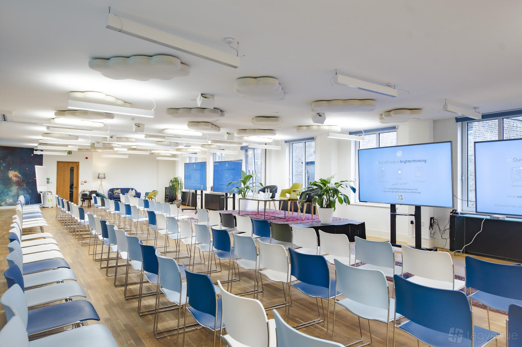 A meeting centre with rows of chairs, floating cloud lights, large windows, and multiple screens at Wallacespace Spitalfields.