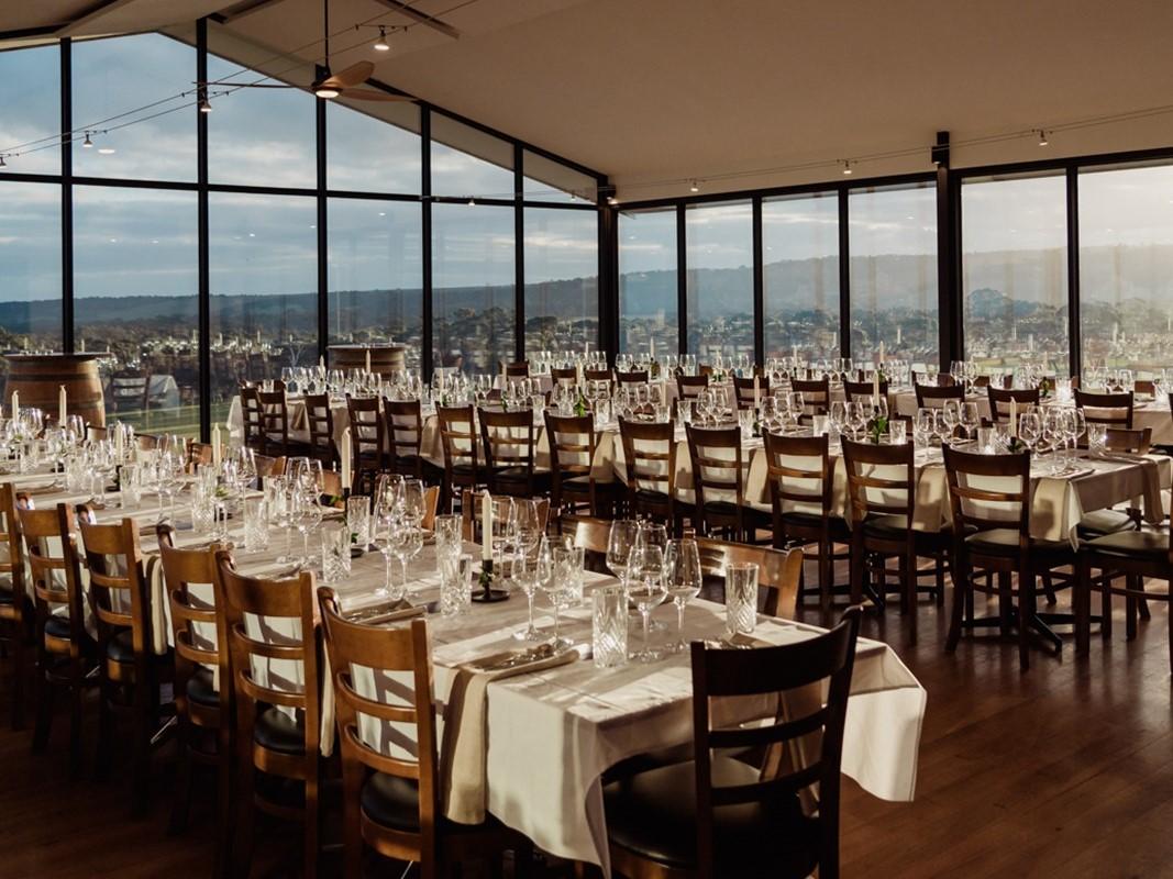 A function room with floor-to-ceiling windows, wooden chairs, and banquet tables set with glassware at The Del Rios Vineyard and Winery.