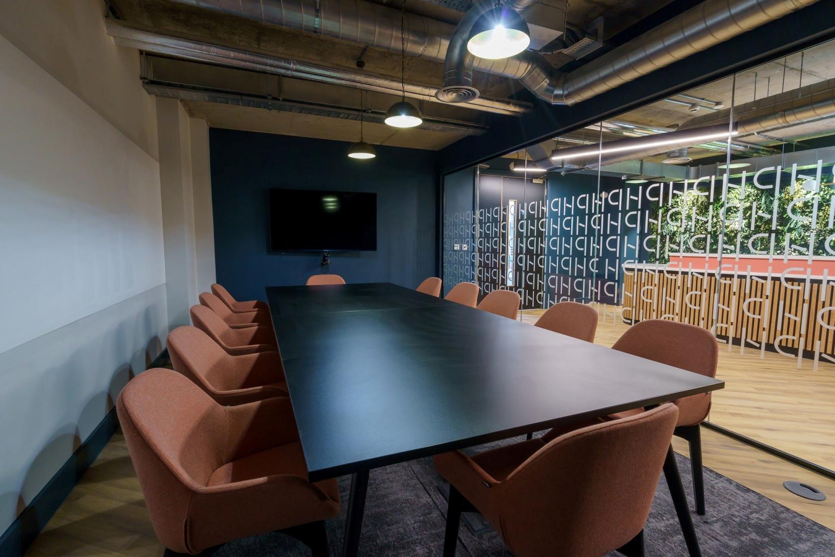 A meeting room with a long black conference table, rust-colored chairs, and a wall-mounted monitor at Wizu Workspace - Newcastle - Portland House.