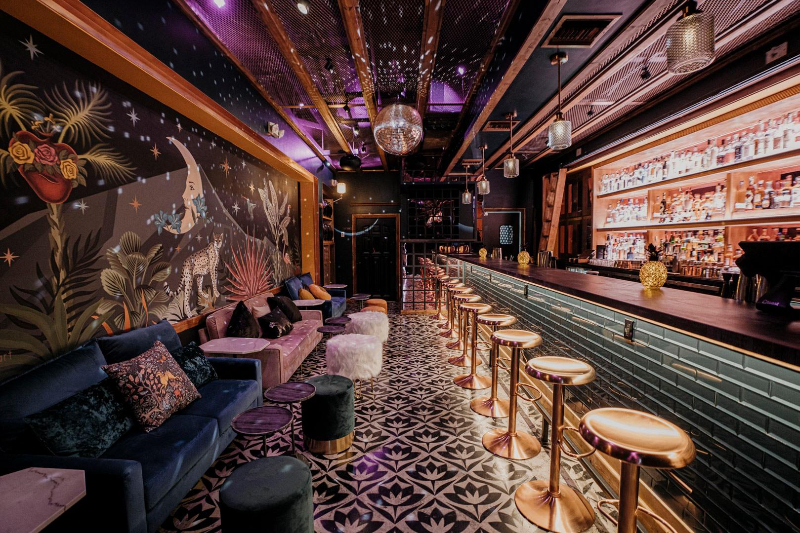 A cocktail bar with a mural wall, velvet seating, geometric tile floor, and backlit bar at La Yolanda Mezcalería Bar.