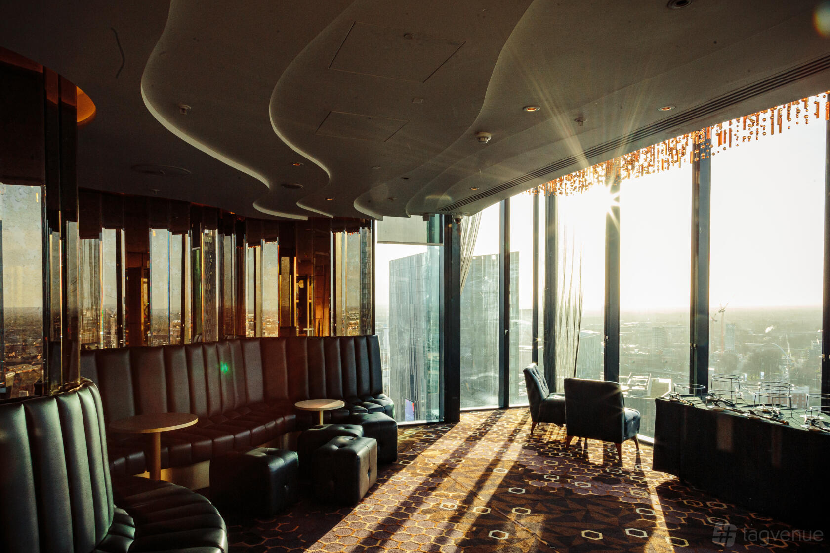 A lounge in a bar with floor-to-ceiling windows, city skyline views, curved banquette seating, and patterned carpet at Cloud 23.