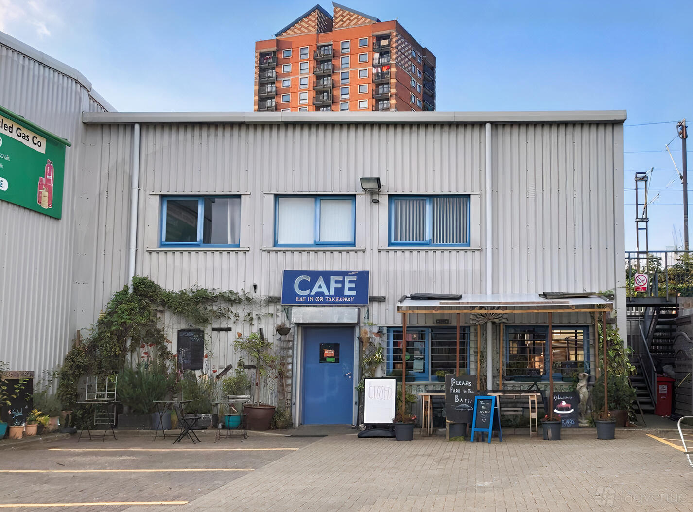 A café with a corrugated metal exterior, potted plants, and blue signage at Old Station Yard Cafe.