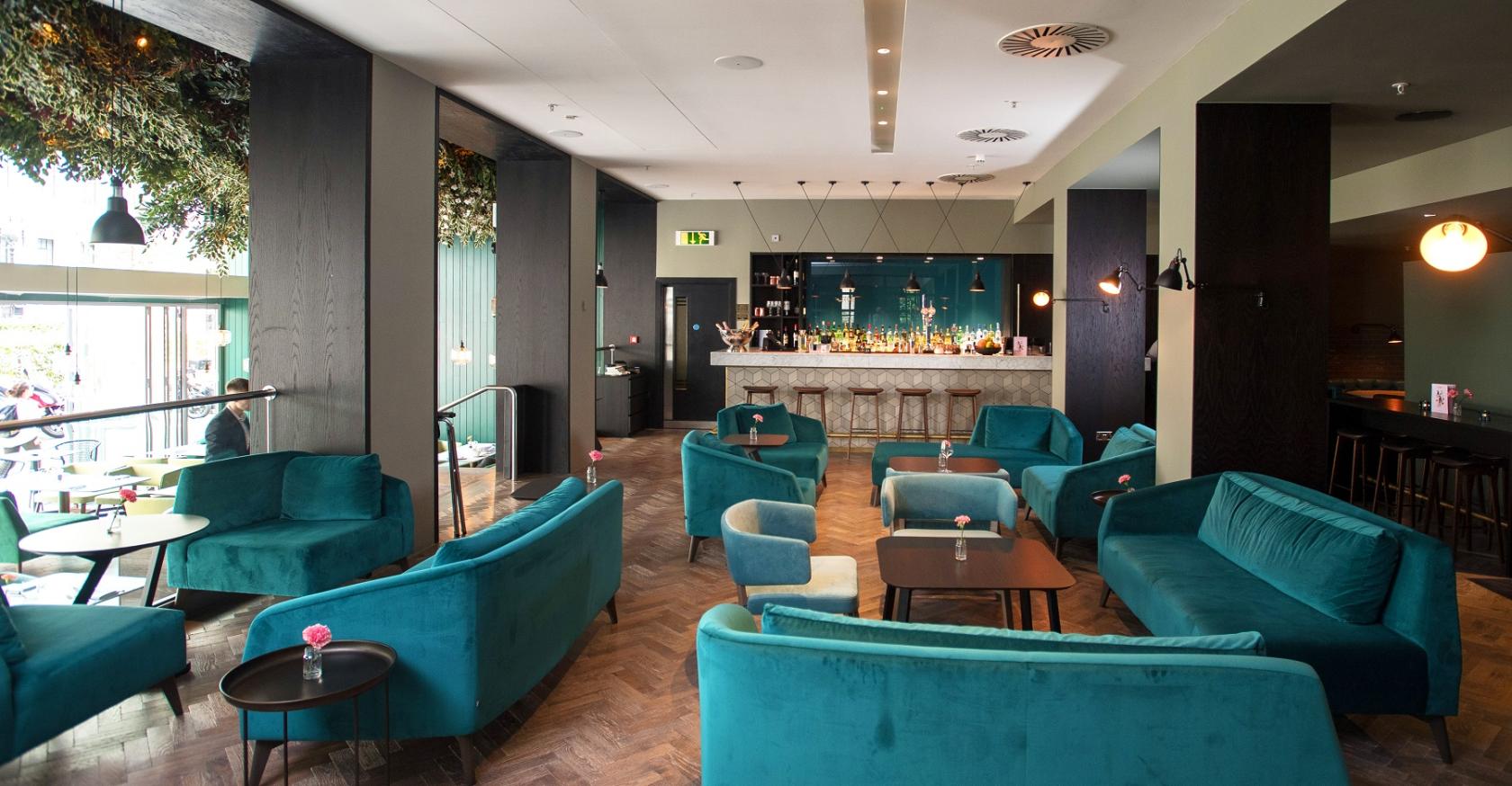 A hotel bar with teal velvet sofas, wooden flooring, and a marble-topped bar at Apex City of London.