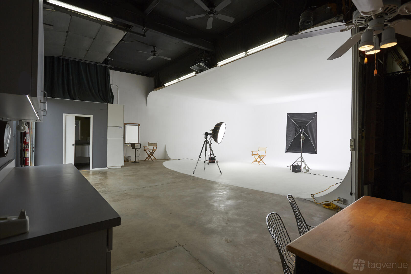 A photo studio with a large white cyclorama wall, studio lighting, director’s chairs, and concrete floors at 3-Wall Cyc with Ceiling.