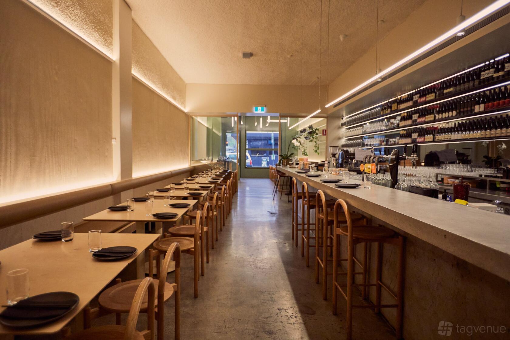 A restaurant dining room with wooden tables, matching chairs, and a long bar lined with shelves of wine at Eazy Peazy.