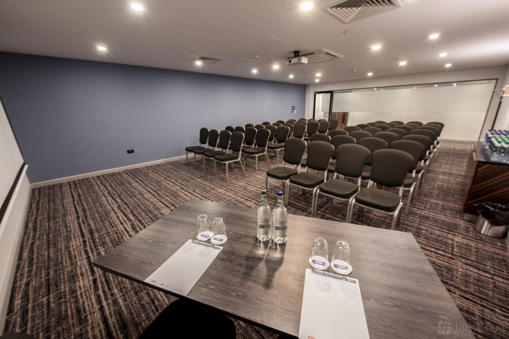 A hotel meeting room with rows of gray chairs, patterned carpeting, and a bottled water setup at The BCEC.