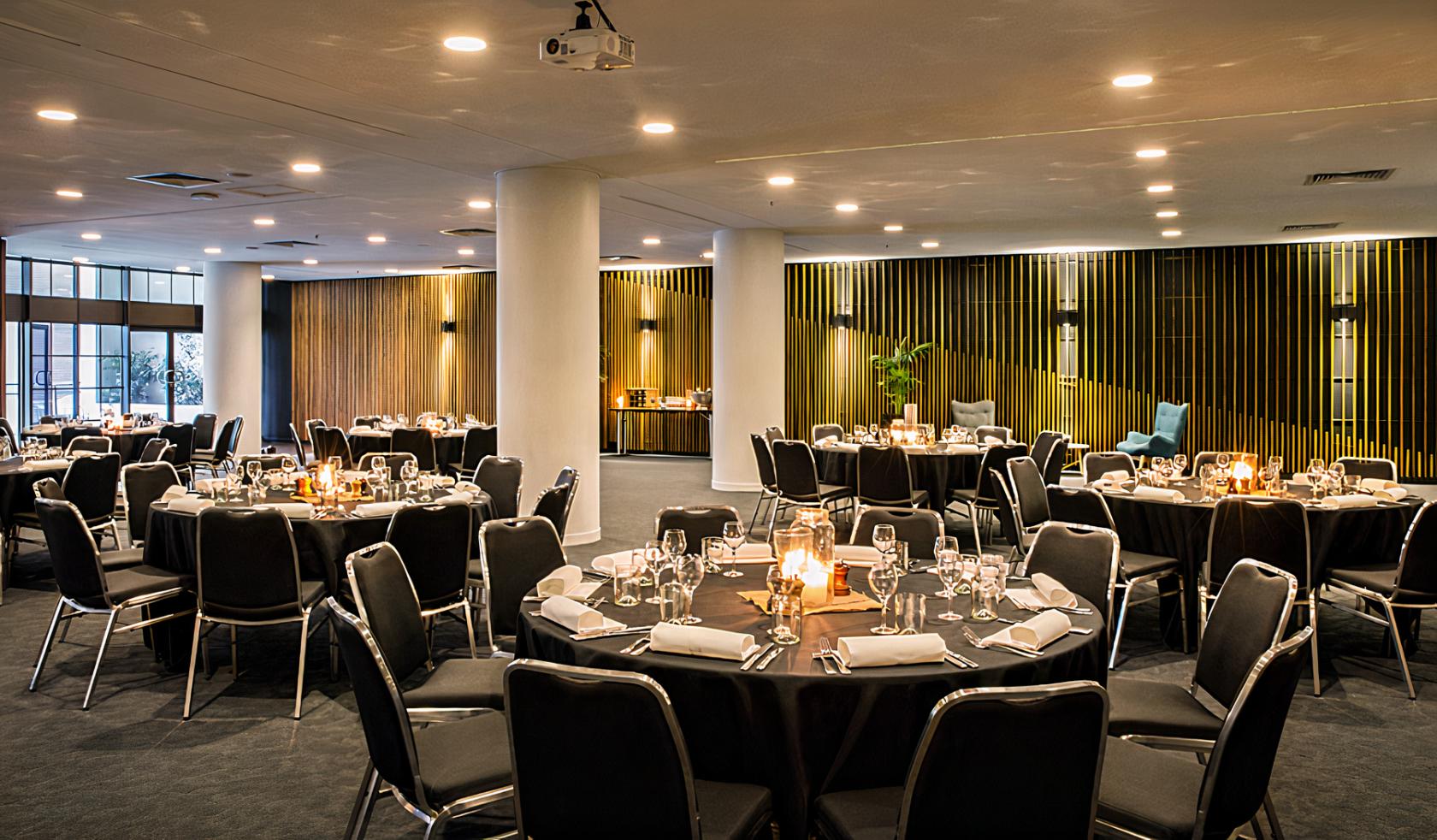 An event space with round tables set with glassware, black chairs, and warm lighting at Ibis Melbourne Hotel and Apartments.