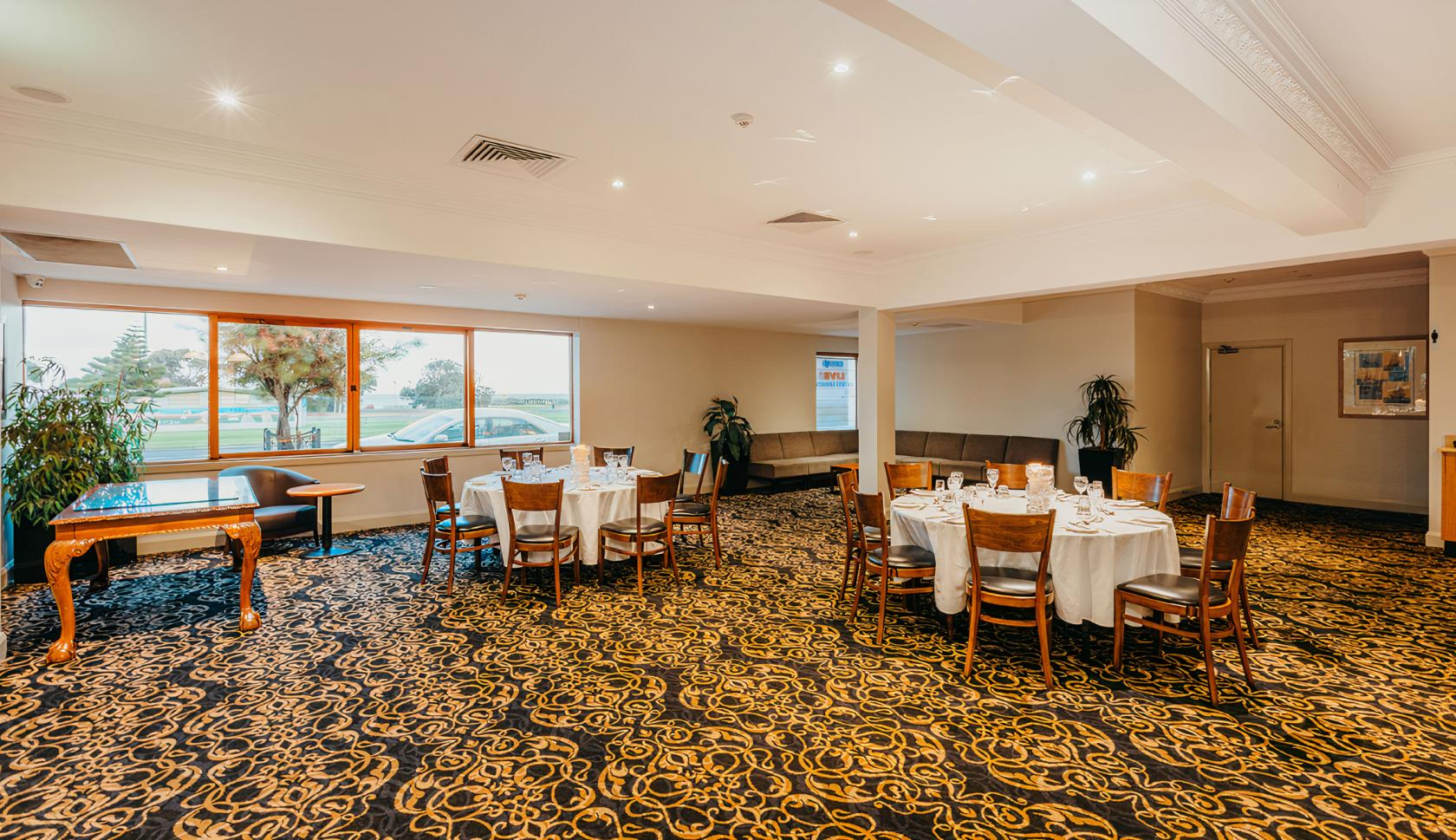 An event space with round tables, white linens, patterned carpet, and large windows overlooking greenery at Rye Hotel.
