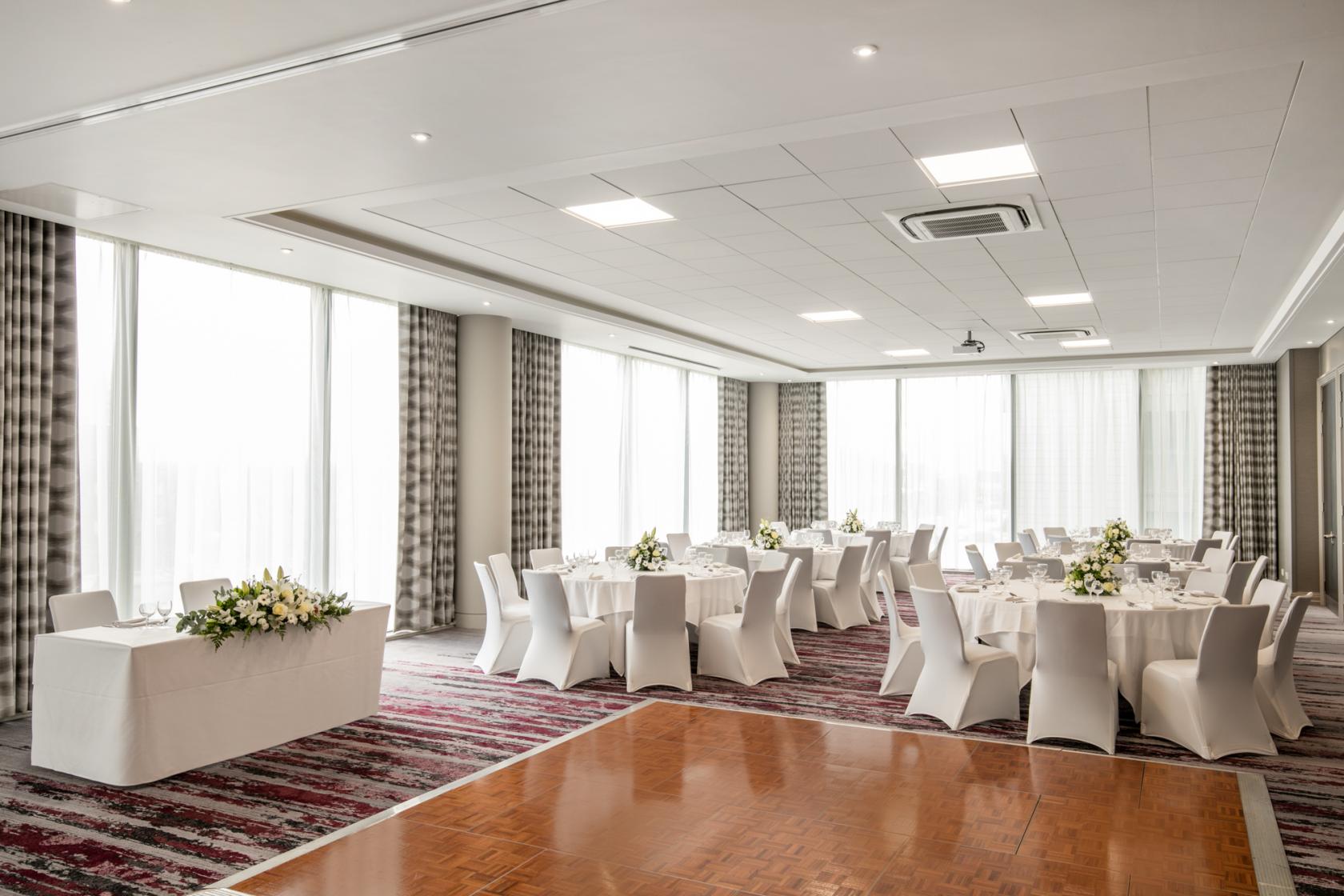 An event space with round tables, white chairs, floral centerpieces, and large windows at Crowne Plaza London Docklands