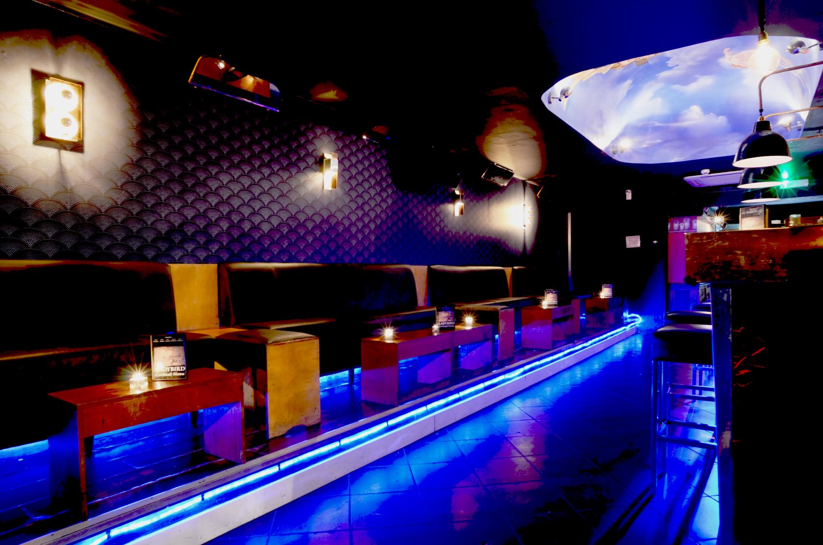 An event space in a cocktail bar with black tufted walls, low wooden tables, and blue LED floor lighting at The Ladybird Bar.