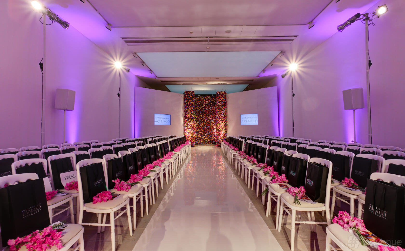 A gallery hall with rows of white chairs, a glossy runway, purple uplighting, and a flower wall at Saatchi Gallery.