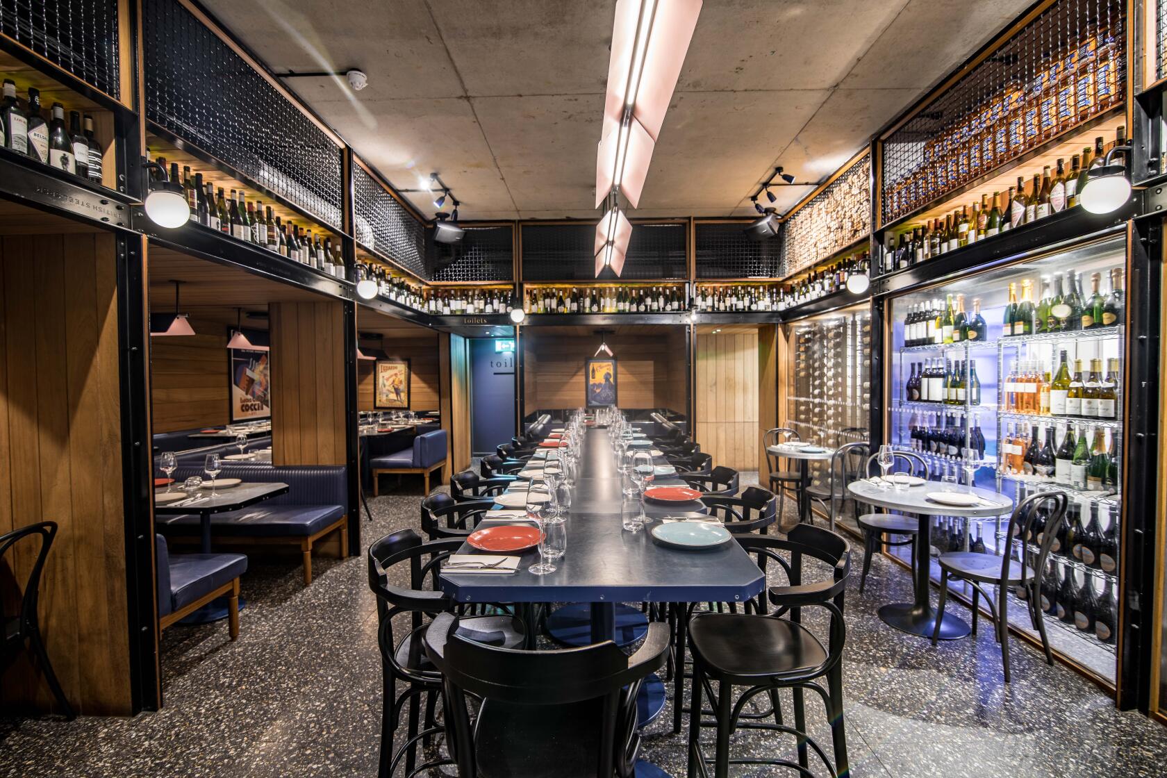 A dining room with a long communal table, black chairs, wine bottle displays, and blue banquette seating at temper Covent Garden.